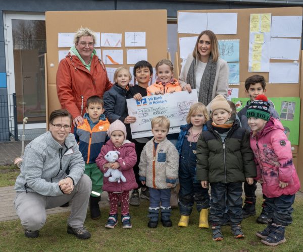 Eine Gruppe von Kindern und Erziehern steht vor einer mit vielen Bildern behängten Pinnwand und hält einen großen Scheck.
